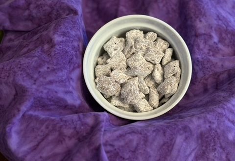A small white ramekin full of Muddy Buddy snack mix lays atop a dark purple cloth napkin, draped as a background. Photo by Diana Ludwick, copyright 2026.