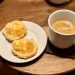 A small white plate holds two halves of a freshly toasted buttermilk biscuit, slathered with Irish butter and French orange marmelade. A fresh mug of coffee sits at the right of the frame.