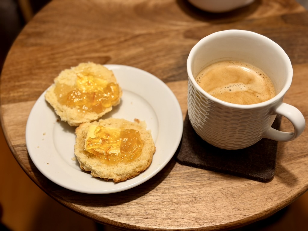 A small white plate holds two halves of a freshly toasted buttermilk biscuit, slathered with Irish butter and French orange marmelade. A fresh mug of coffee sits at the right of the frame.