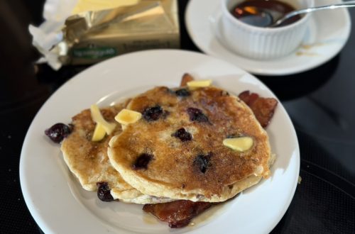 A short stack of blueberry buttermilk pancakes with ample butter and maple syrup on a white plate. a stick of butter in gold foil is in the back left. A ramekin of maple syrup is at the back right.