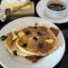 A short stack of blueberry buttermilk pancakes with ample butter and maple syrup on a white plate. a stick of butter in gold foil is in the back left. A ramekin of maple syrup is at the back right.