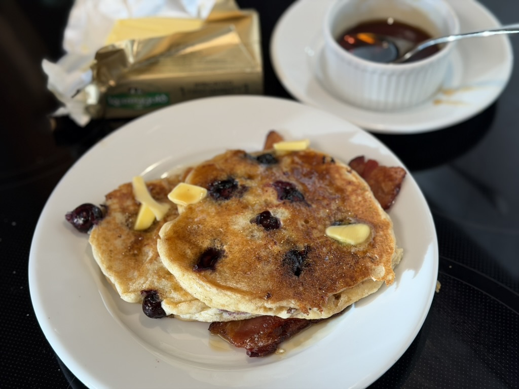 A short stack of blueberry buttermilk pancakes with ample butter and maple syrup on a white plate. a stick of butter in gold foil is in the back left. A ramekin of maple syrup is at the back right.