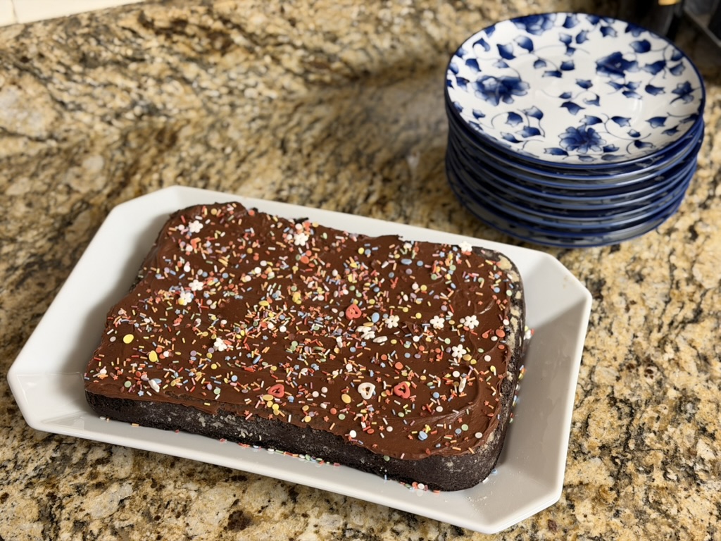 A celebration cake - chocolate ganache with chocolate cake, on an octagonal white platter on a granite countertop. A stack of blue and white floral patterned plates sit behind the cake.