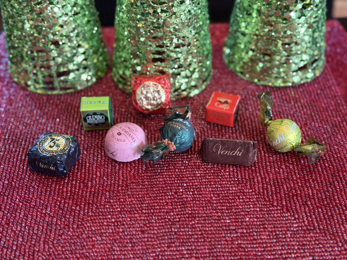 An assortment of brightly colored, foil-wrapped chocolate truffles, bon bons, cubes and batons are strewn on a sparkling glass-beaded placemat. The bases of three green sequined Christmas trees are in the background.
